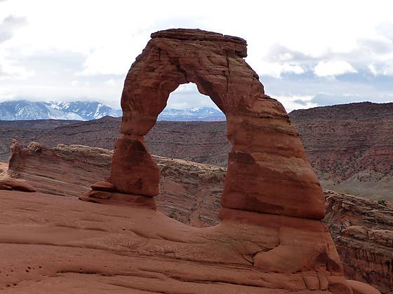 Delicate Arch framed by the La Sal Mountains to the east, partially hidden by clouds.